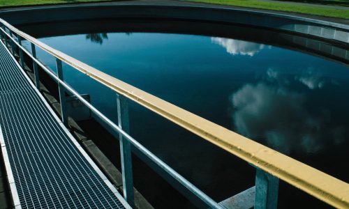 Grated walkway with a yellow-topped railing beside dark, still water that reflects clouds.